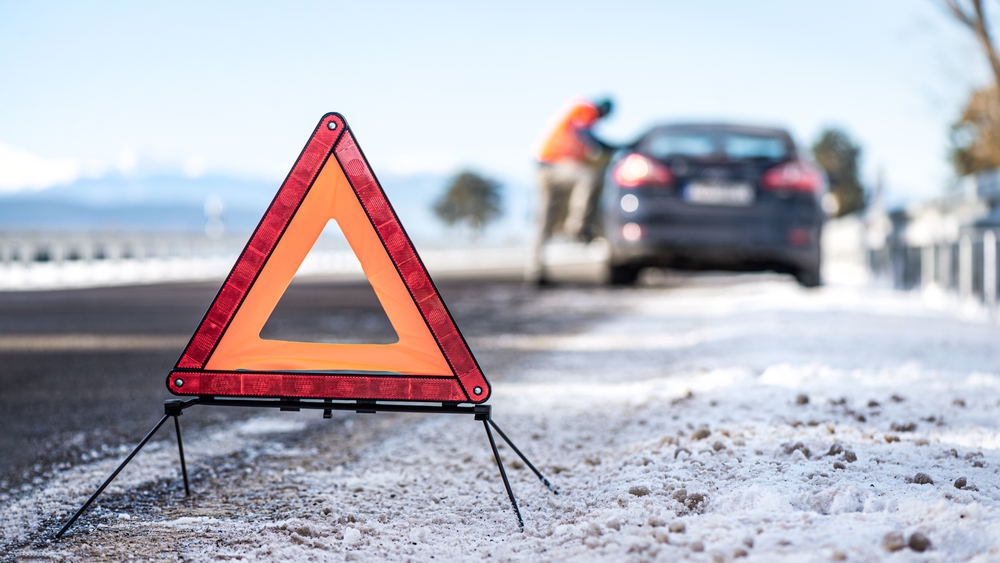 An orange emergency warning triangle on a snowy roadside after a black ice crash in New Jersey, with a driver inspecting vehicle damage.