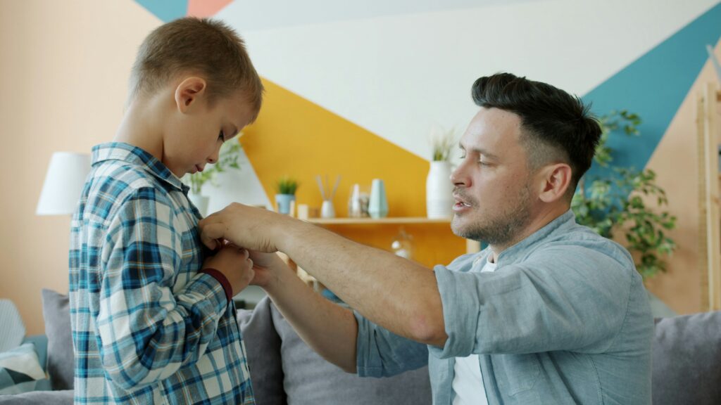 Father helping his young son button his shirt at home, illustrative image representing how a catastrophic injury can suddenly change a parent's role in the family.
